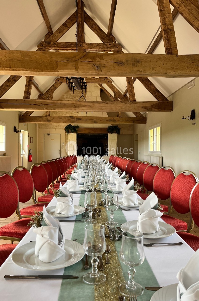 Salle de réception avec une longue table dressée, nappes blanches, serviettes pliées et chaises rouges, sous une charpente…