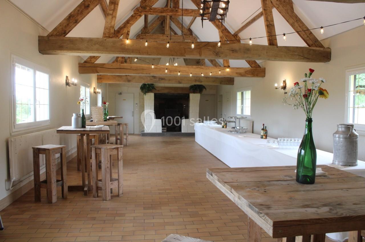 Salle lumineuse avec poutres apparentes, tables en bois, décorations florales et buffet installé au fond.