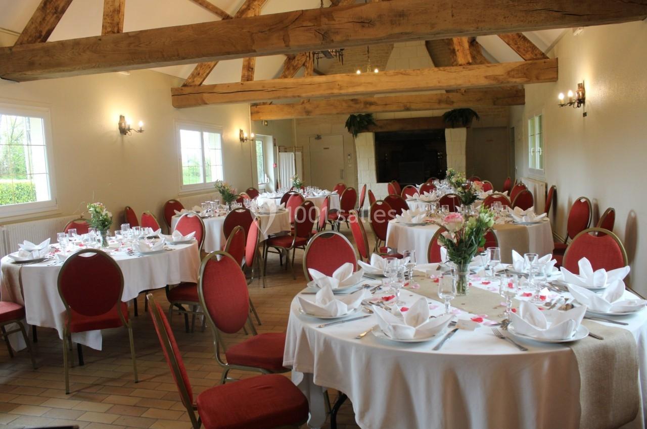 Salle de réception avec tables rondes dressées, nappes blanches, chaises rouges et poutres apparentes au plafond.