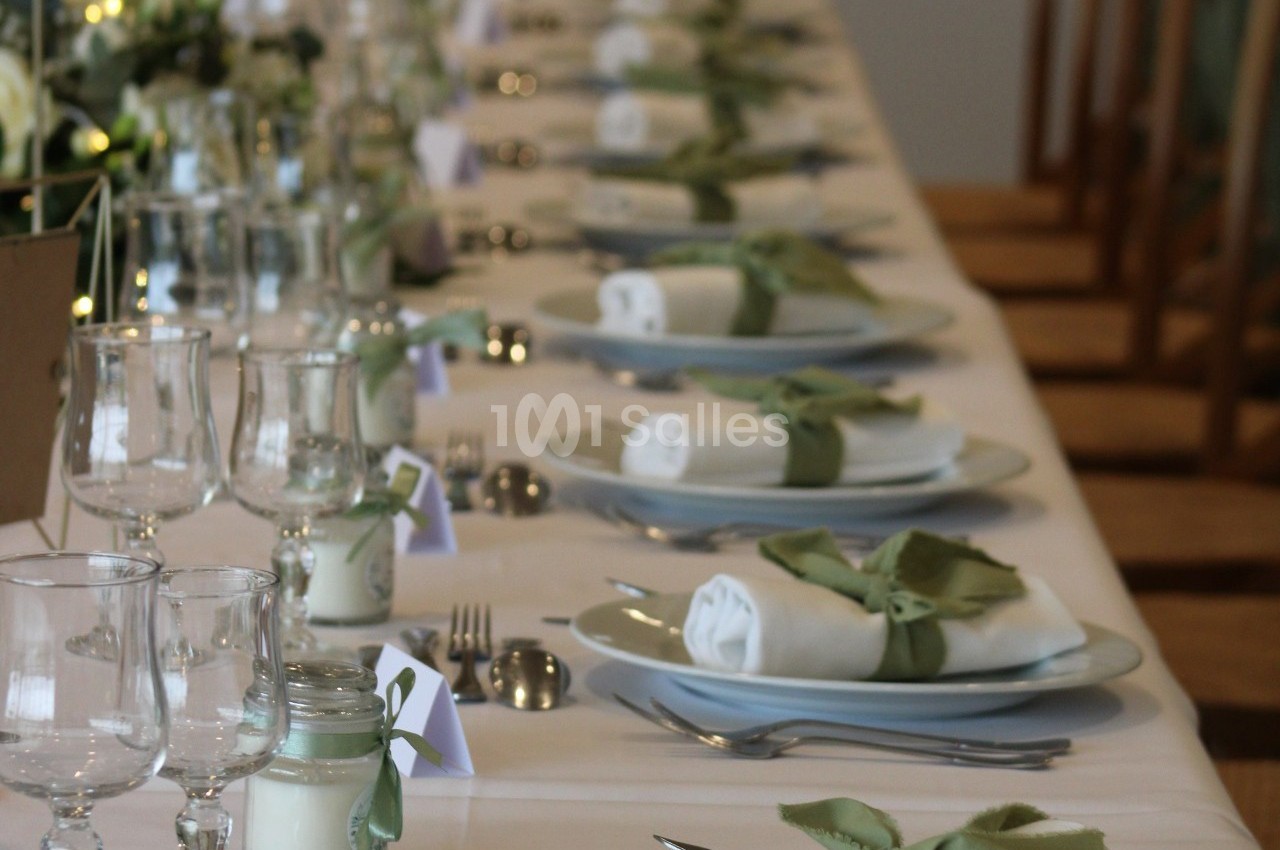 Table dressée avec nappes blanches, serviettes roulées entourées de rubans verts, verres et décorations florales.