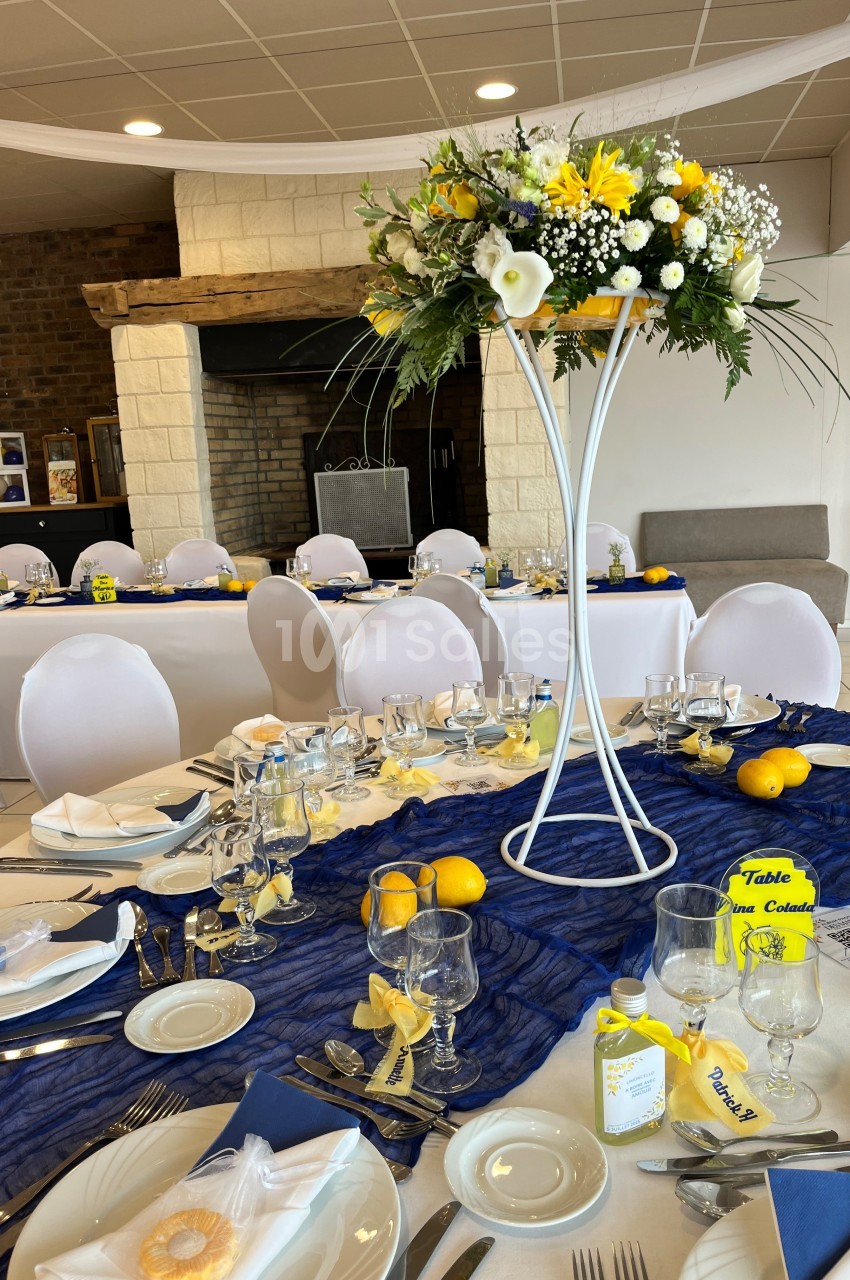 Salle de réception décorée avec des tables dressées, nappes bleues, centres de table fleuris et détails jaunes.