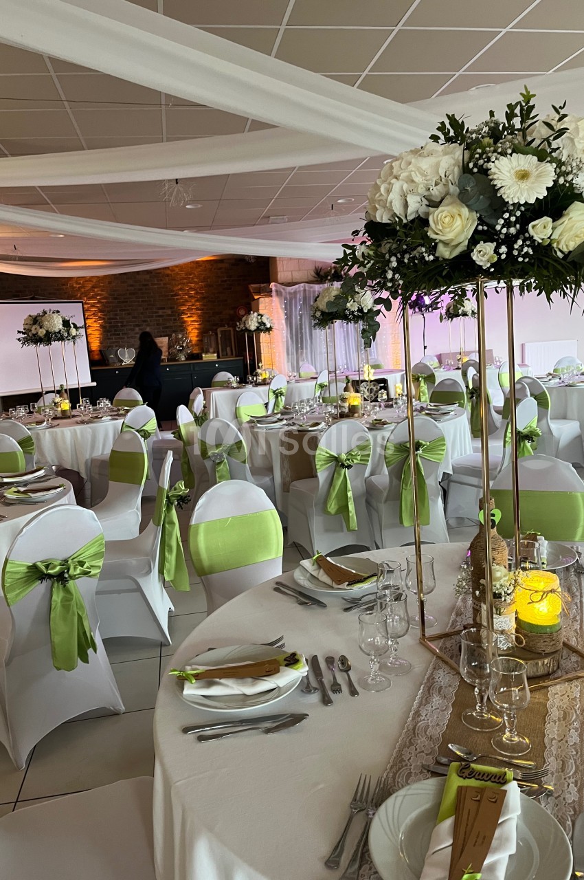 Salle de réception décorée pour un mariage, avec tables dressées, chaises ornées de rubans verts et compositions florales.