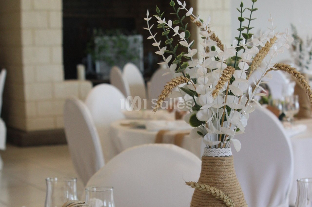 Vase en corde avec des fleurs blanches et des épis de blé, posé sur une table dans une salle de réception élégante.