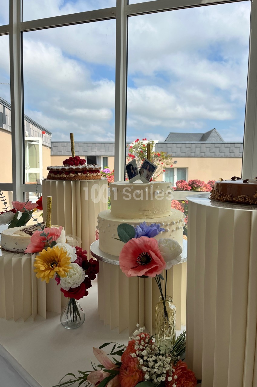 Gâteaux décorés de fleurs et bougies, exposés sur des supports devant une fenêtre avec vue sur des bâtiments et le ciel.