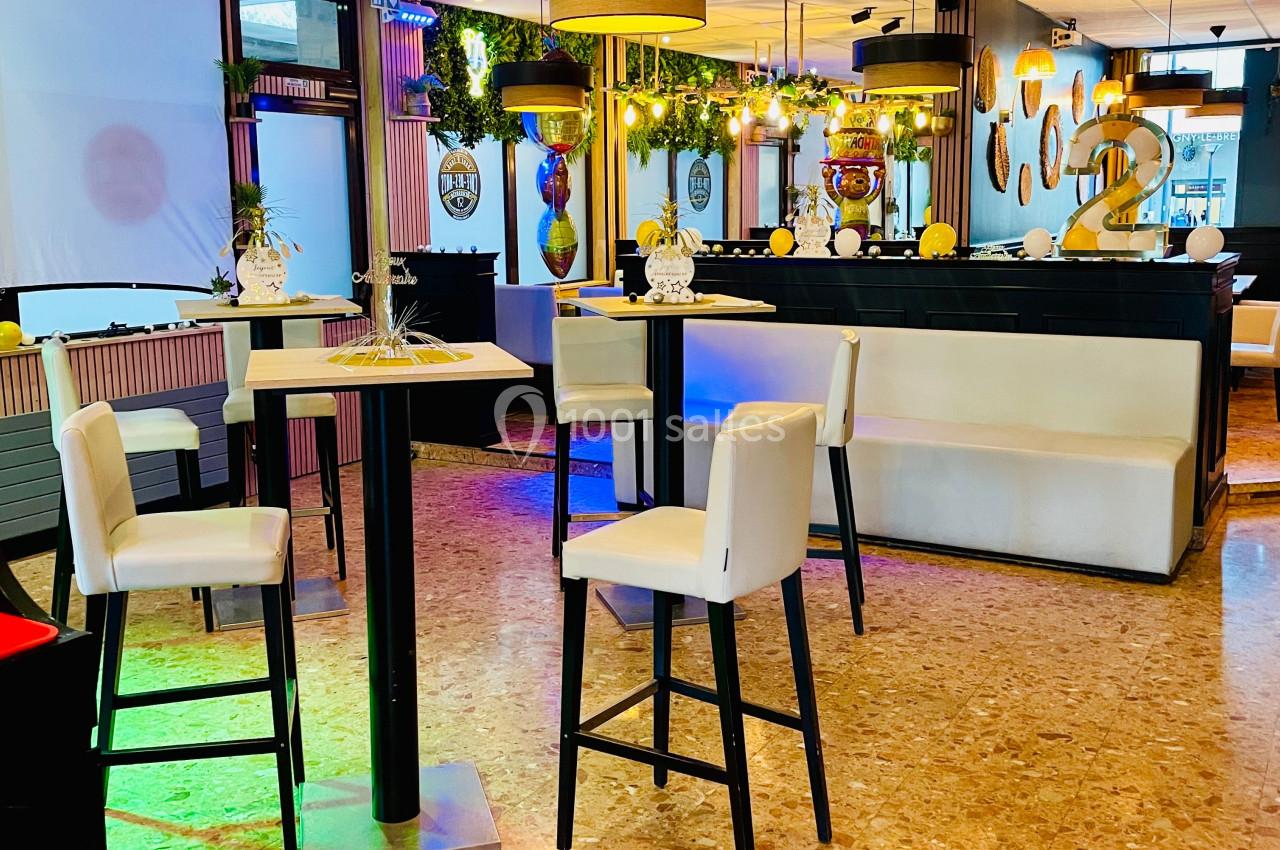 Salle lumineuse avec tables hautes, chaises blanches, décorations modernes et plantes suspendues.