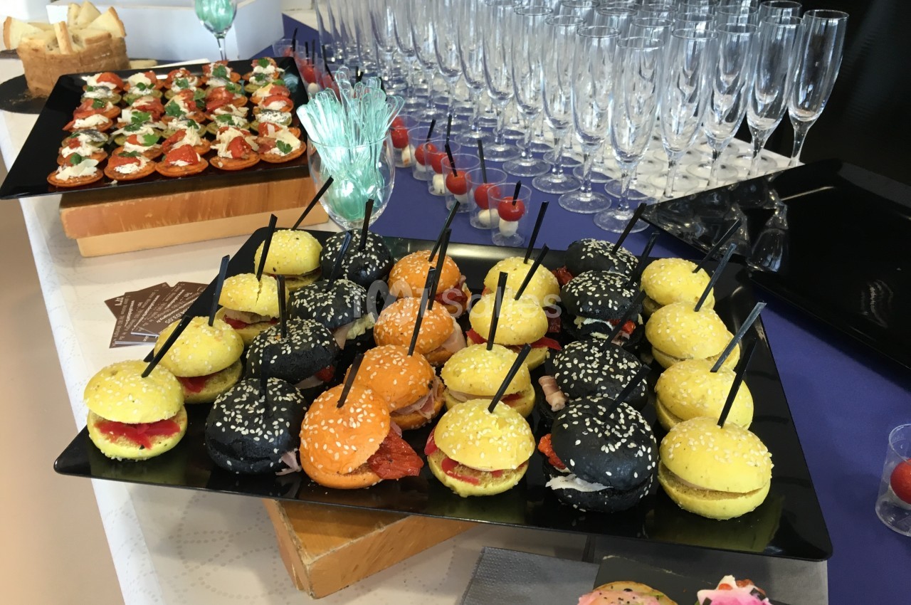 Plateaux de mini-burgers colorés et amuse-bouches disposés sur une table avec des flûtes à champagne alignées.
