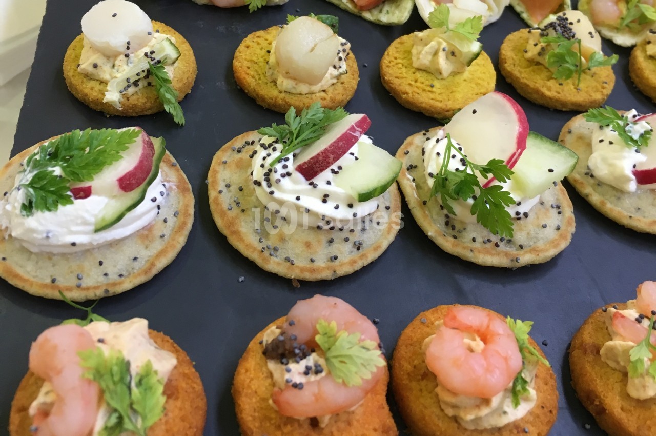 Assortiment de canapés variés garnis de crevettes, légumes frais, fromage et herbes sur un plateau noir.