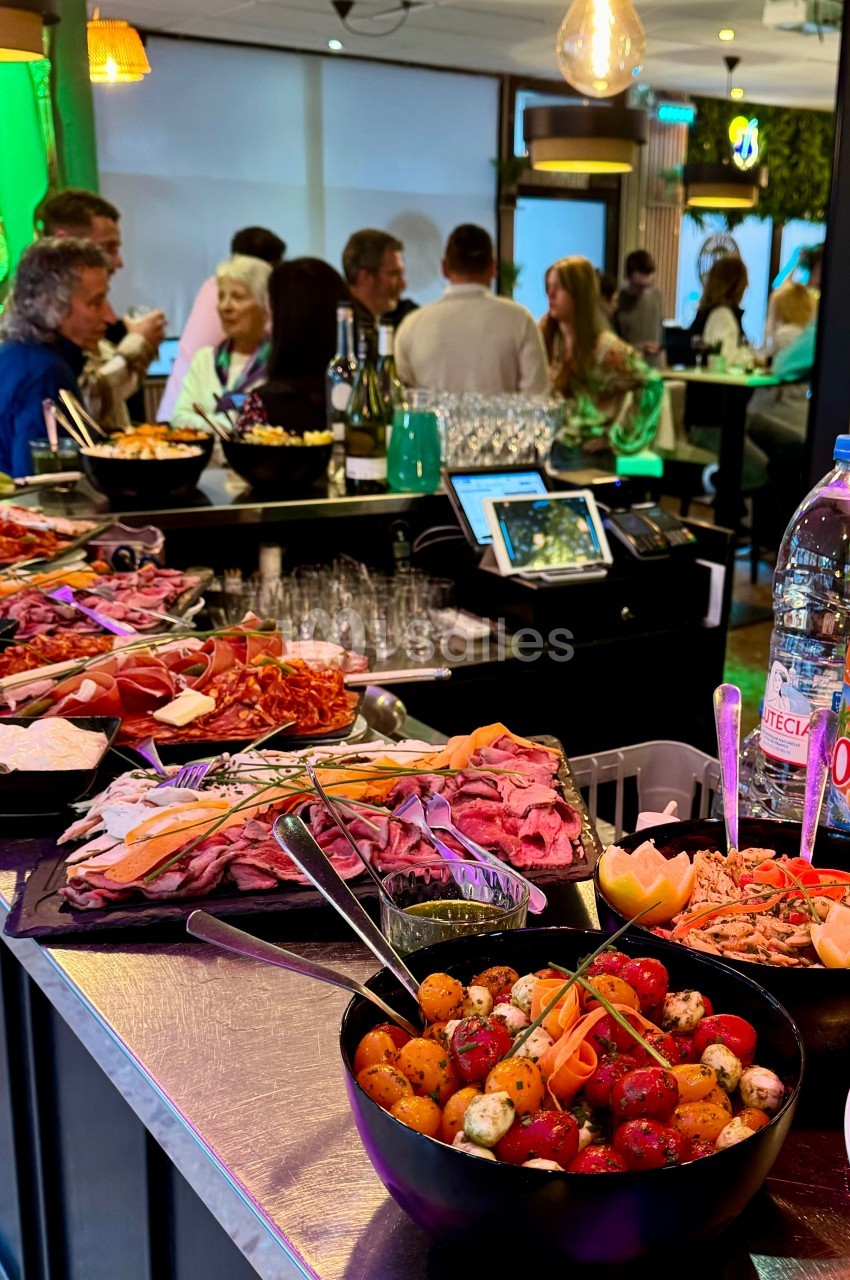 Buffet varié avec charcuterie, légumes marinés et boissons, dans un bar animé avec des convives en arrière-plan.
