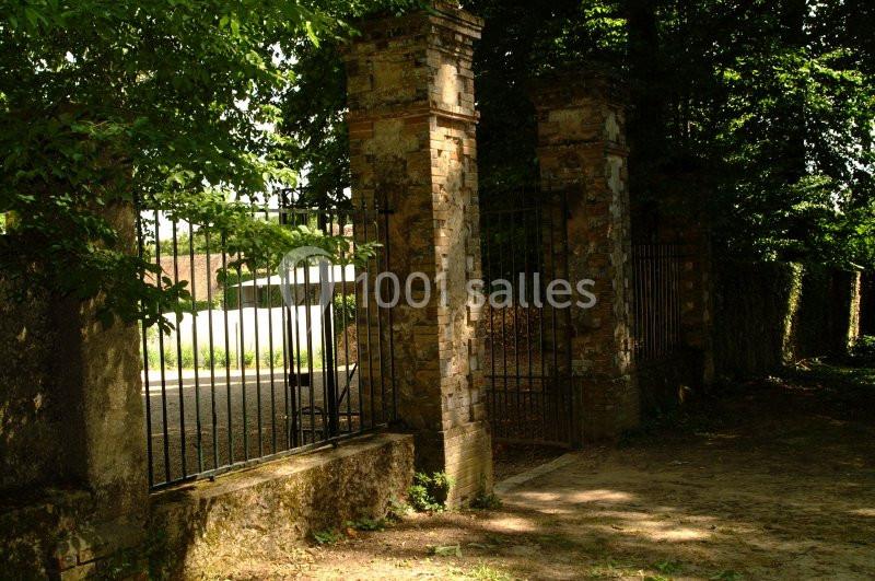 Portail en fer forgé encadré par des piliers en pierre, entouré de végétation et donnant sur une allée ensoleillée.