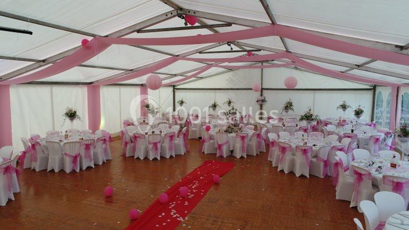 Salle de réception décorée en rose et blanc avec des tables rondes, des chaises ornées de rubans et un tapis rouge central.
