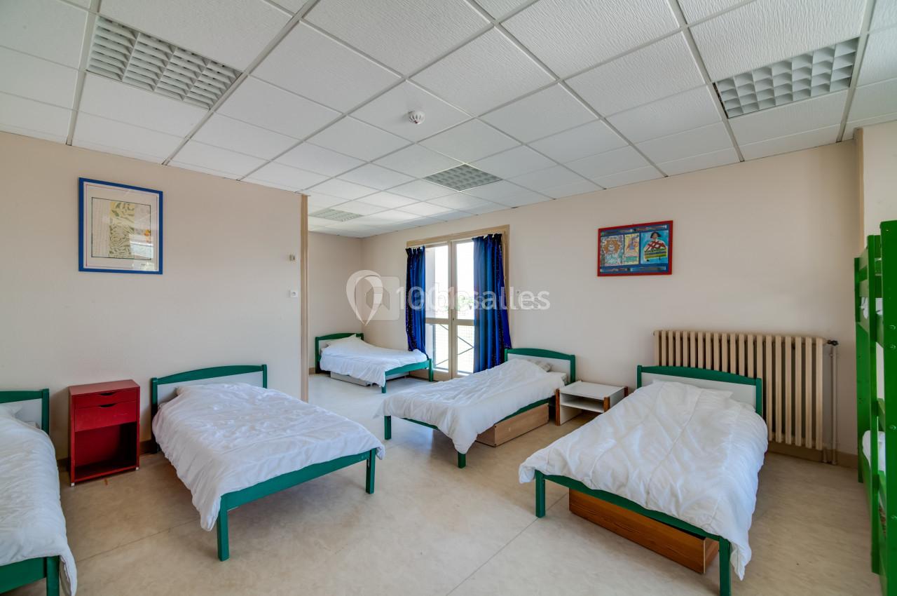 Dortoir lumineux avec cinq lits simples, tables de chevet rouges et fenêtres donnant sur l'extérieur.