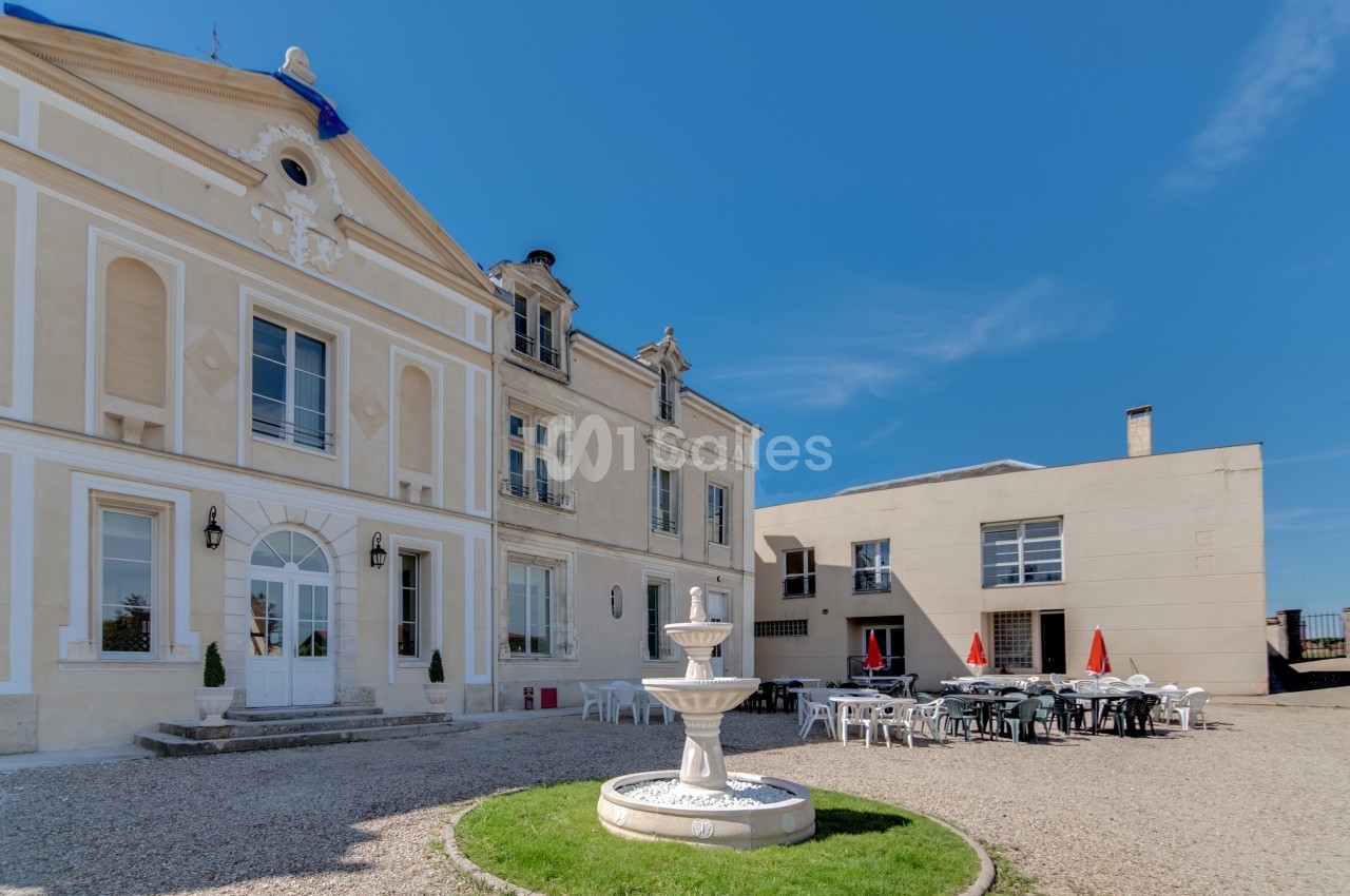 Façade d'un bâtiment historique avec une fontaine au premier plan et une terrasse aménagée sous un ciel dégagé.