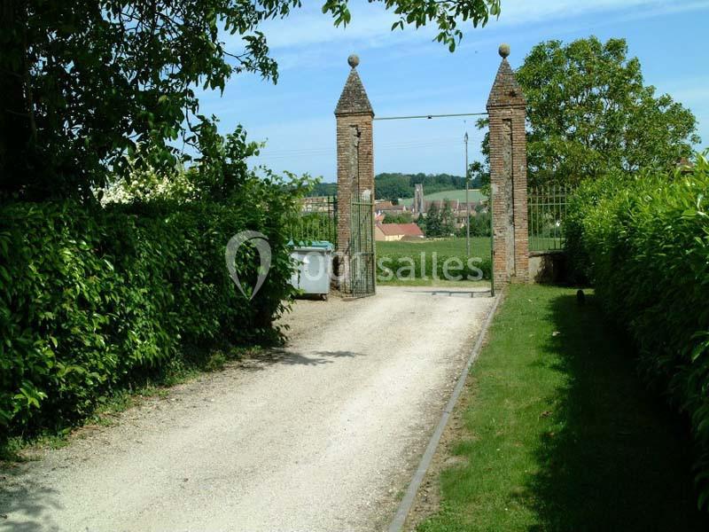 Allée en gravier bordée de haies menant à un portail en briques avec vue sur un village à l'arrière-plan.