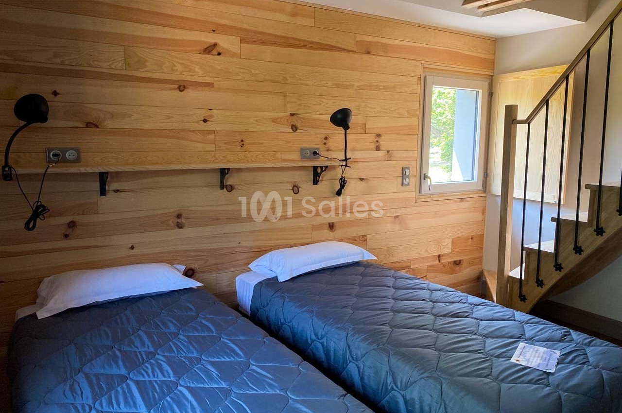 Deux lits simples avec couvre-lits gris dans une chambre aux murs en bois, éclairée par une fenêtre et un escalier visible.