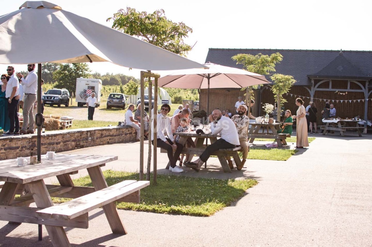 Personnes assises à des tables en bois sous des parasols dans une cour ensoleillée près d'un bâtiment en pierre.