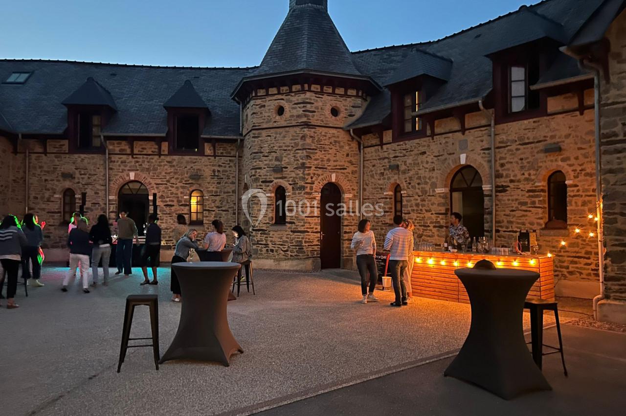 Cour intérieure d’un bâtiment en pierre, éclairée en soirée, avec des tables hautes et des personnes discutant près d’un bar.