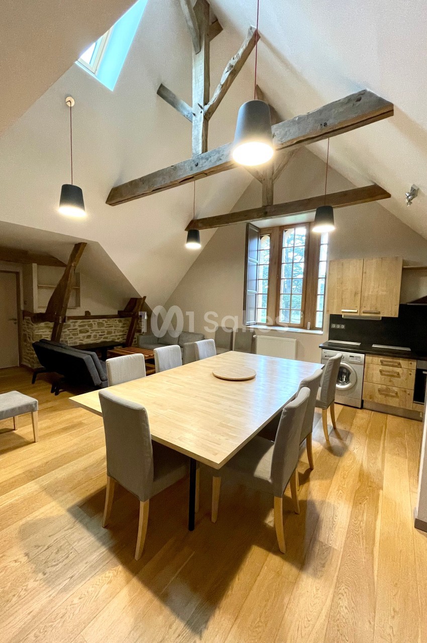 Salle à manger lumineuse avec table en bois, chaises grises, cuisine équipée et poutres apparentes sous un plafond mansardé.