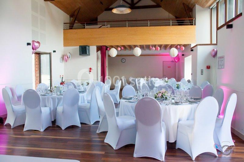 Salle de réception décorée avec des tables rondes, nappes blanches, chaises houssées et éclairage rose.