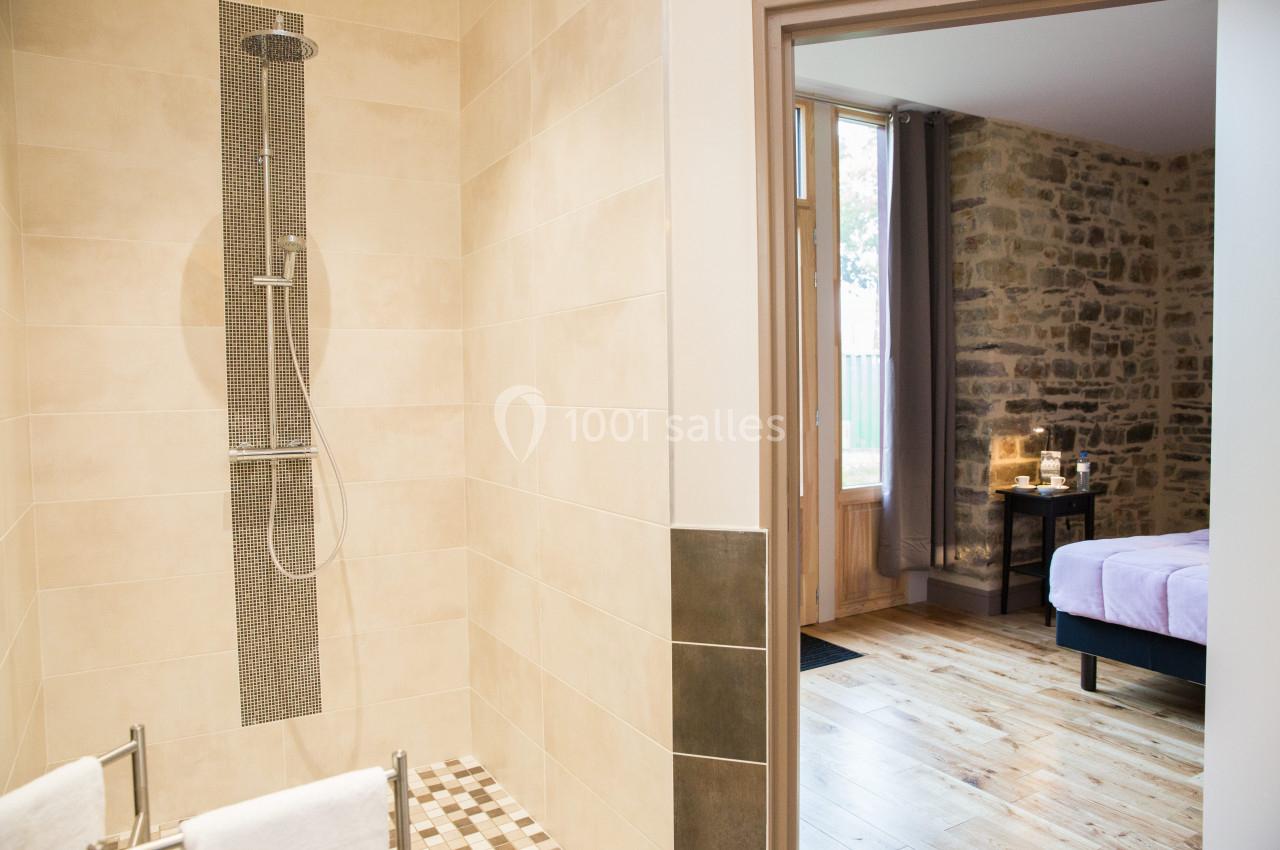 Salle de bain avec douche à l'italienne, vue partielle sur une chambre avec murs en pierre et parquet en bois.