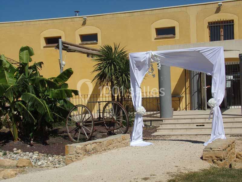Arche de mariage blanche décorée de fleurs, installée devant un bâtiment jaune avec des plantes et une roue en bois.