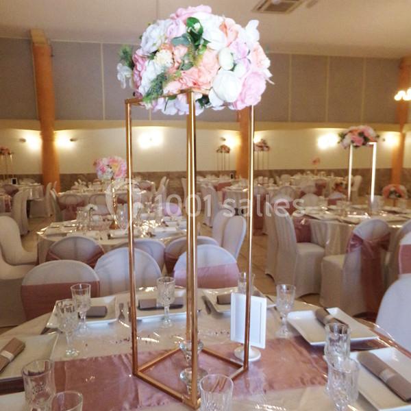 Centre de table floral avec des roses roses et blanches sur un support doré, dans une salle de réception décorée.