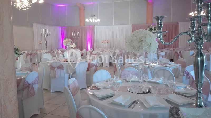 Salle de réception décorée avec des nappes blanches, des chaises ornées de rubans roses et des centres de table fleuris.