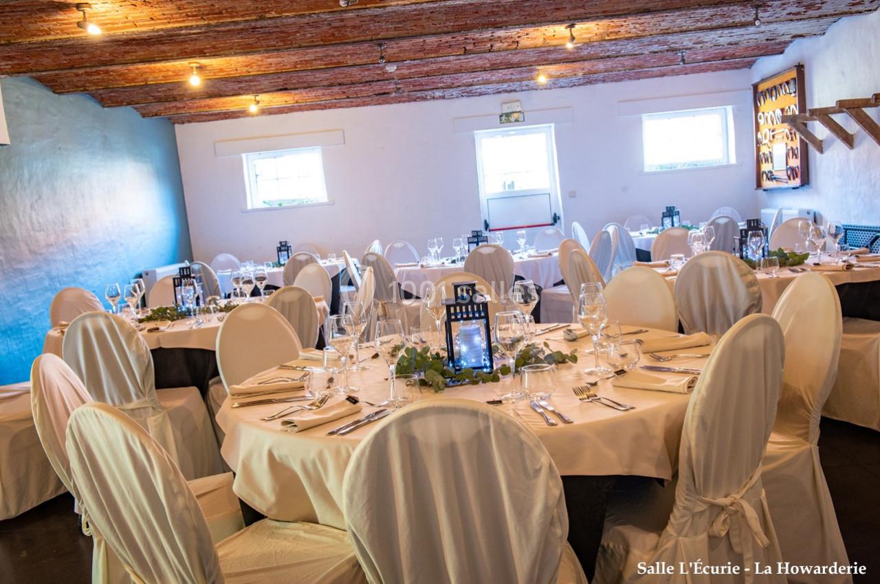 Salle décorée avec des tables rondes, nappes blanches, chaises couvertes et lumière naturelle entrant par les fenêtres.