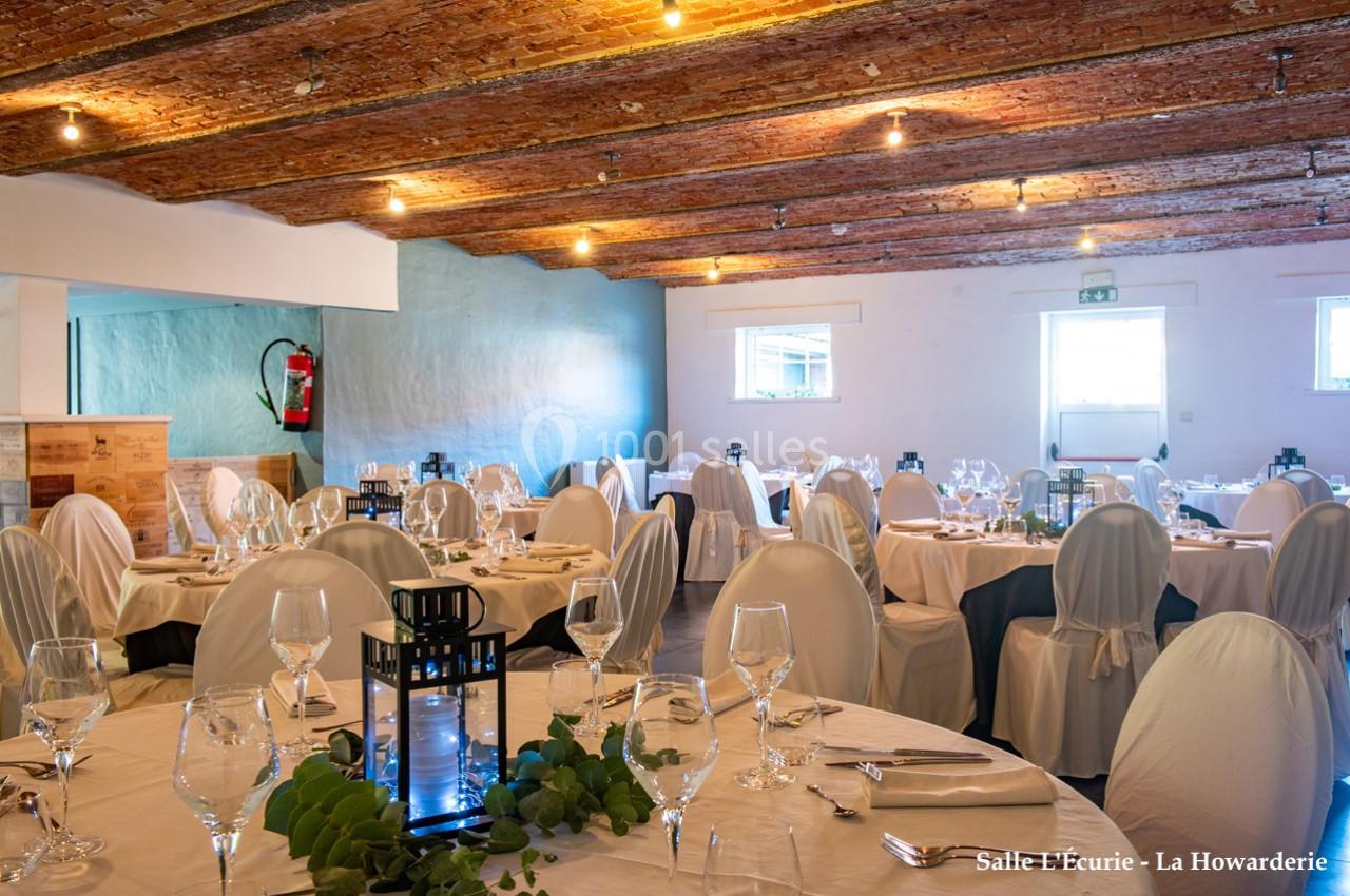 Salle de réception avec tables dressées, nappes blanches, chaises couvertes et plafond en briques apparentes.