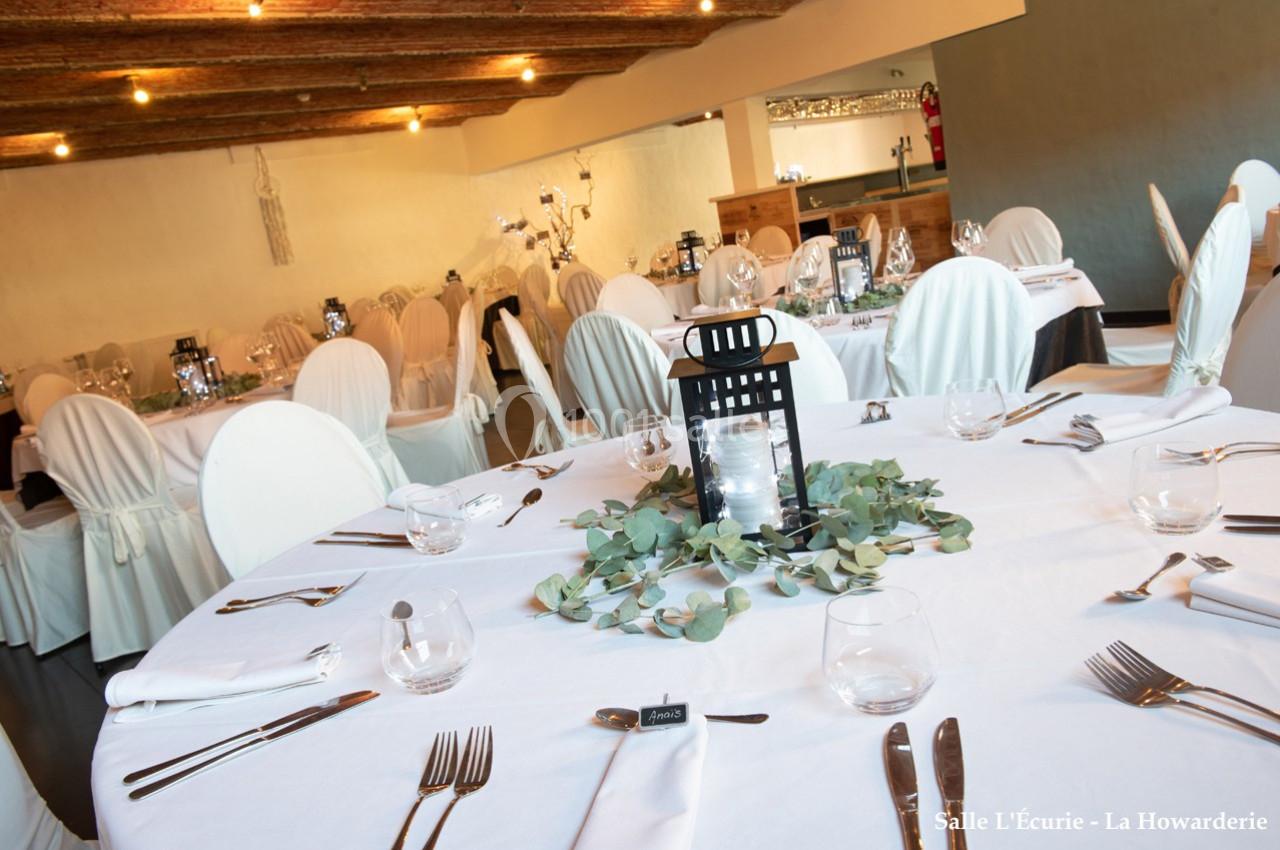 Salle de réception avec tables rondes dressées, nappes blanches, chaises houssées et décoration végétale centrale.