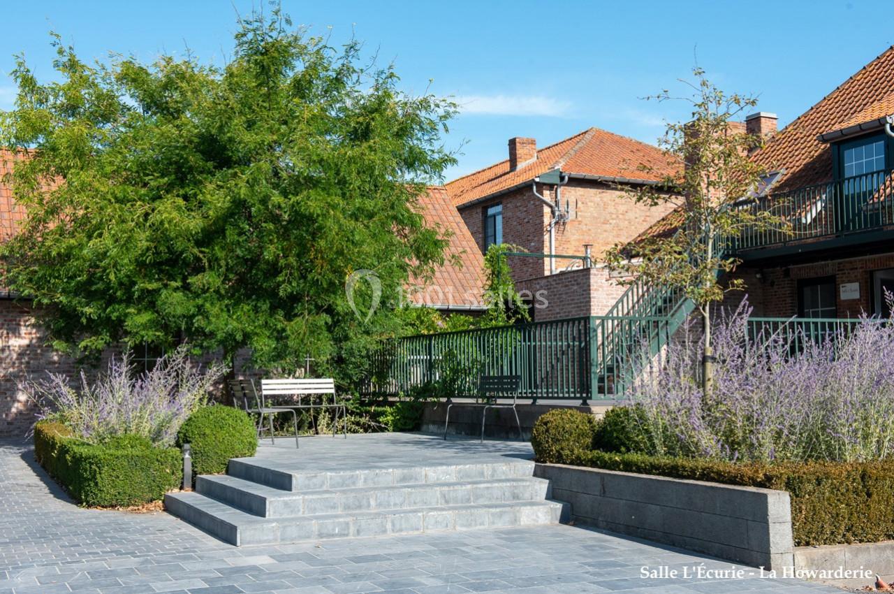 Cour pavée avec escaliers, bancs, arbustes taillés et bâtiments en briques rouges sous un ciel dégagé.