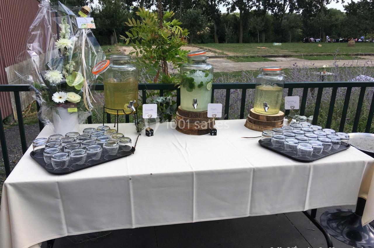 Table avec distributeurs de boissons, verres alignés et décorations florales, en extérieur près d'un espace vert.