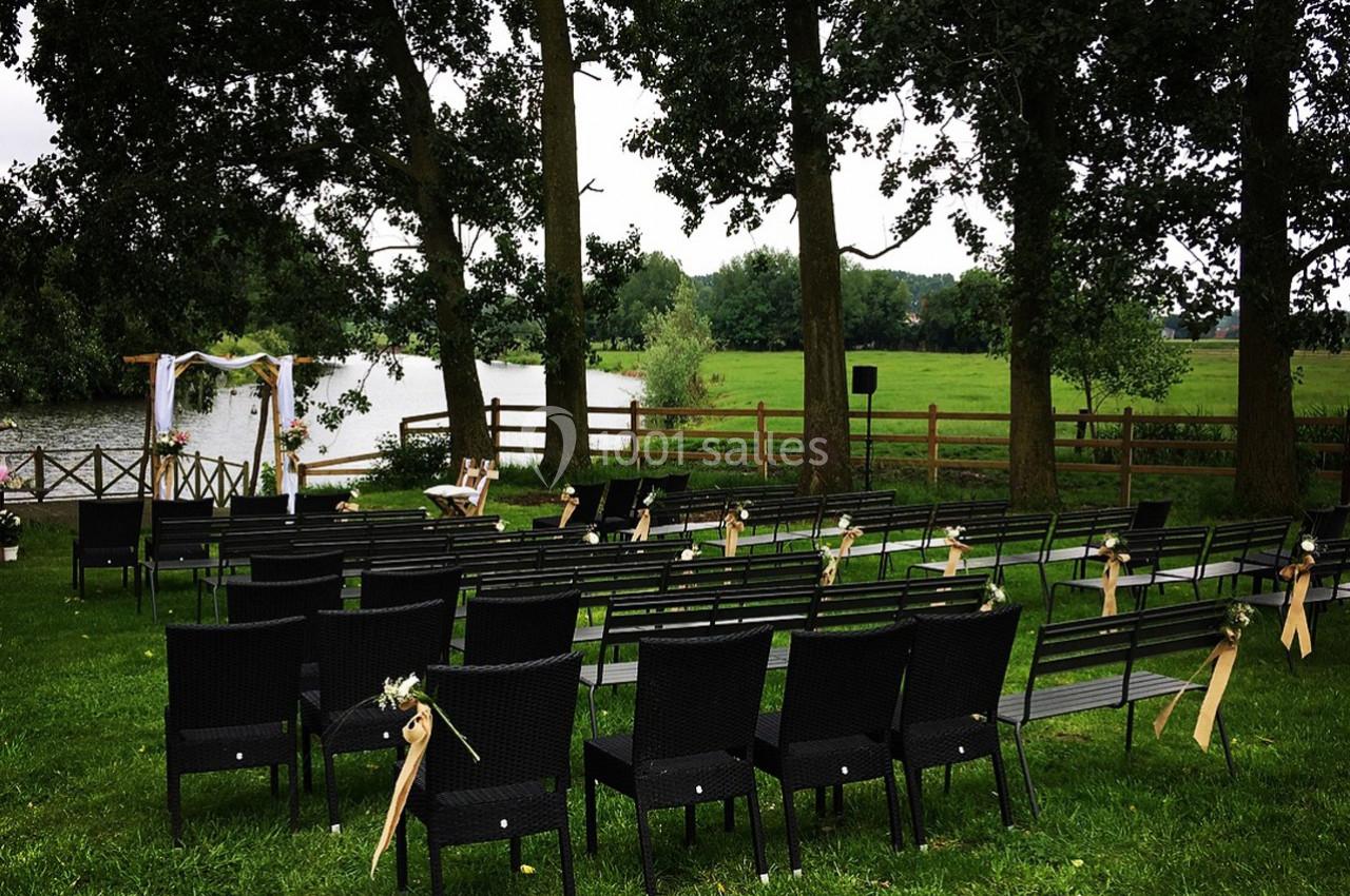 Chaises noires disposées en rangées sur une pelouse, face à une arche décorée près d'un étang entouré d'arbres.