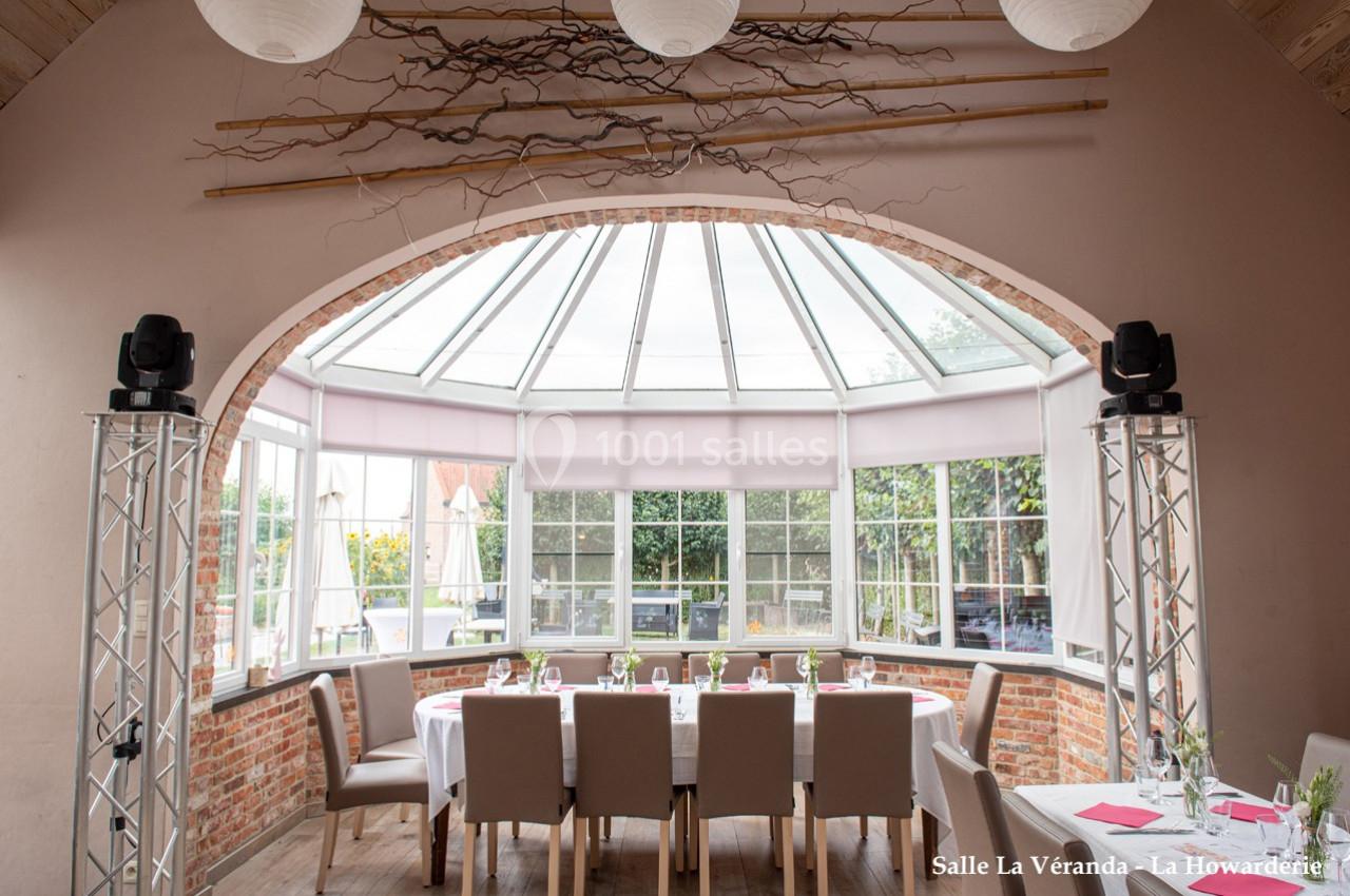 Salle lumineuse avec verrière, table dressée pour un repas, chaises beige clair et décoration naturelle en branches.