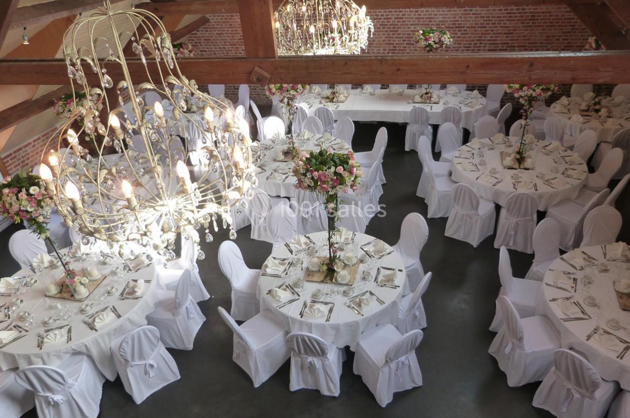 Salle de réception avec tables rondes dressées, nappes blanches, chaises houssées et lustres imposants.