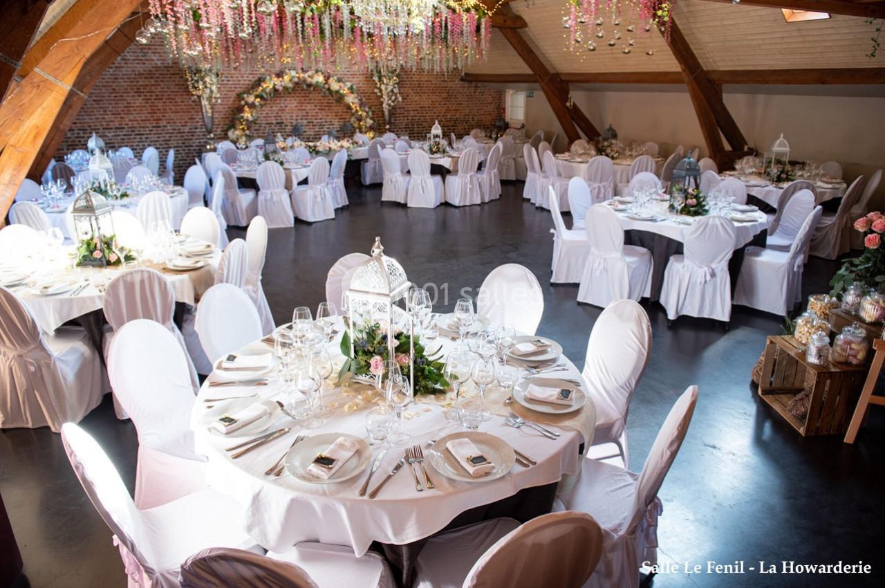 Salle de réception décorée pour un mariage, avec tables rondes dressées, chaises blanches et guirlandes florales suspendues.