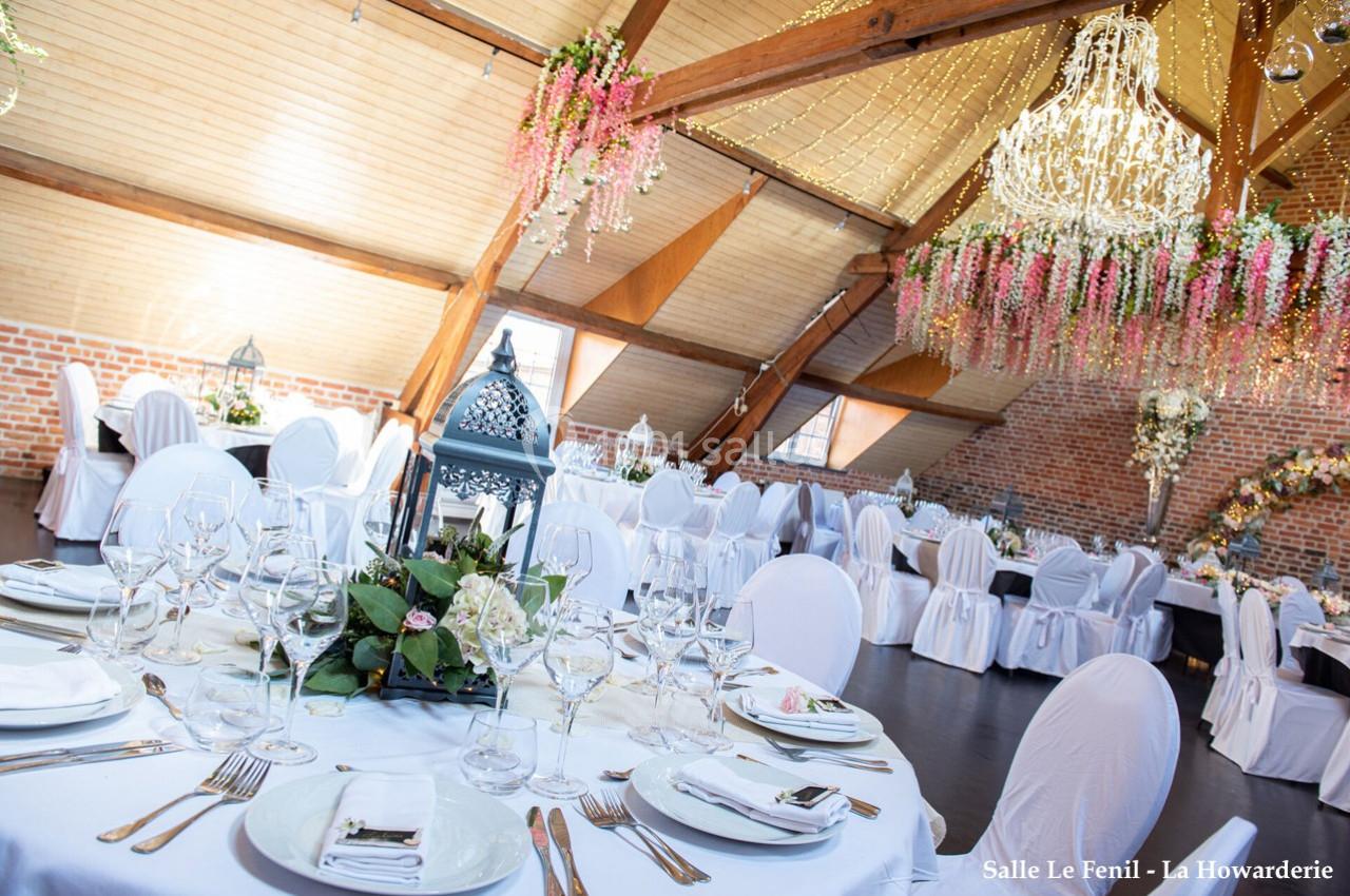 Salle de réception lumineuse avec tables décorées de nappes blanches, fleurs suspendues et lanternes au centre.