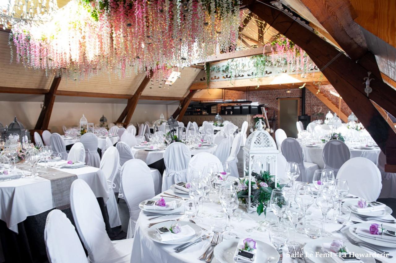 Salle de réception décorée avec des tables rondes, nappes blanches, chaises houssées et guirlandes florales suspendues.
