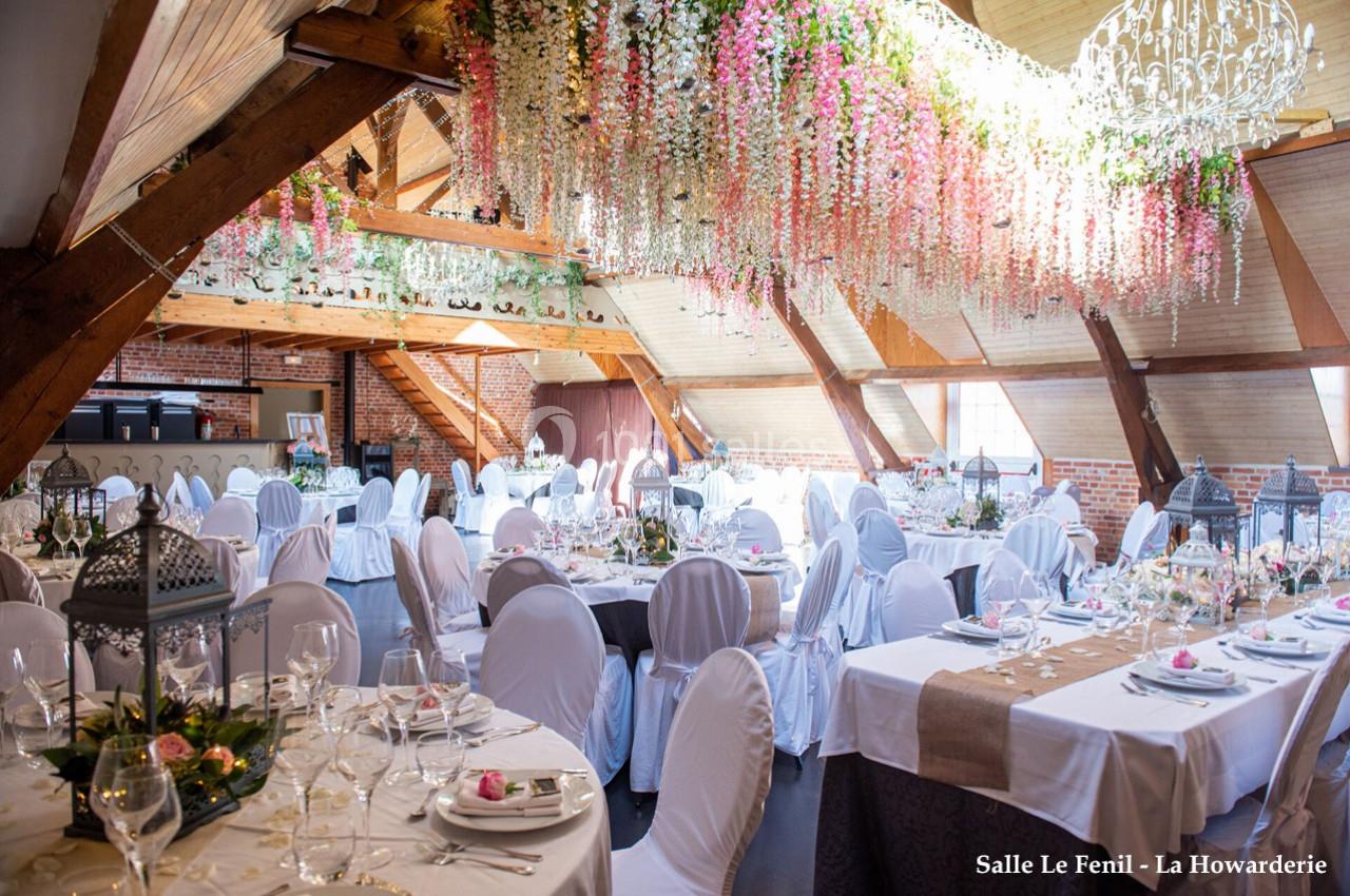 Salle de réception lumineuse avec tables dressées, chaises blanches et décoration florale suspendue au plafond.