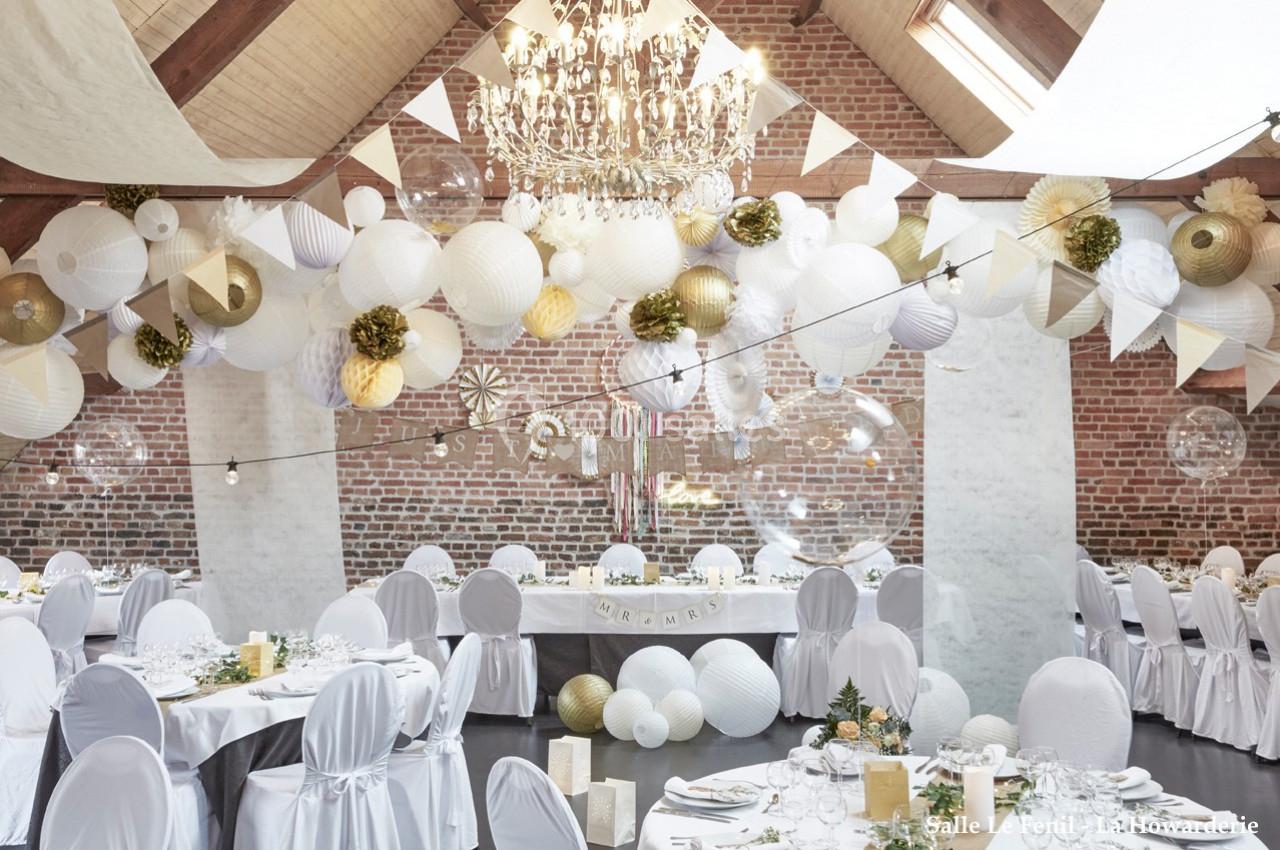 Salle décorée pour un événement avec des lanternes blanches et dorées suspendues, tables dressées et murs en briques…