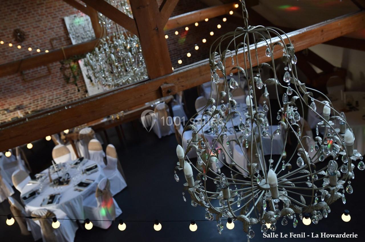 Salle de réception avec tables rondes dressées, chaises blanches et grand lustre en cristal sous une charpente en bois.