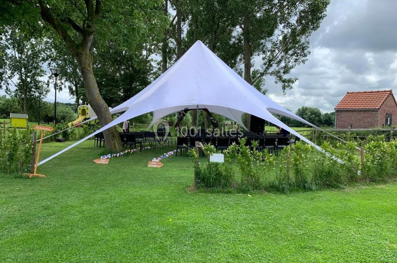 Tente blanche en étoile installée sur une pelouse entourée d'arbres, avec des tables et chaises disposées dessous.