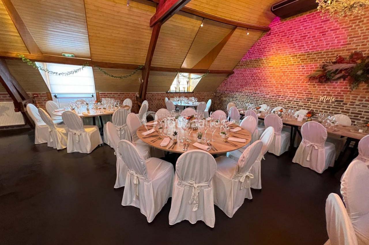 Salle de réception avec tables rondes dressées, nappes blanches et décoration murale en briques et feuillage.