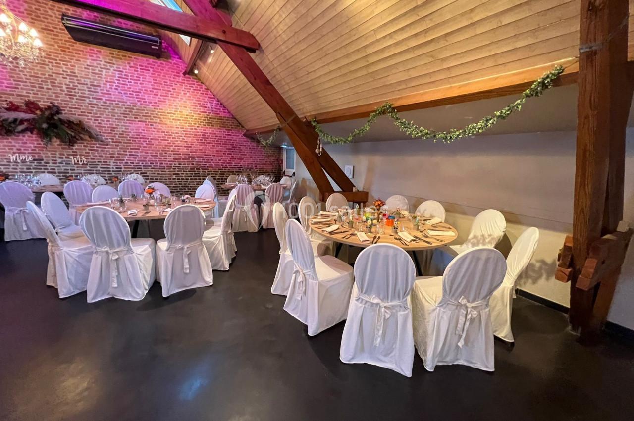 Salle de réception avec tables rondes dressées, chaises blanches et décoration florale sous un plafond en bois.