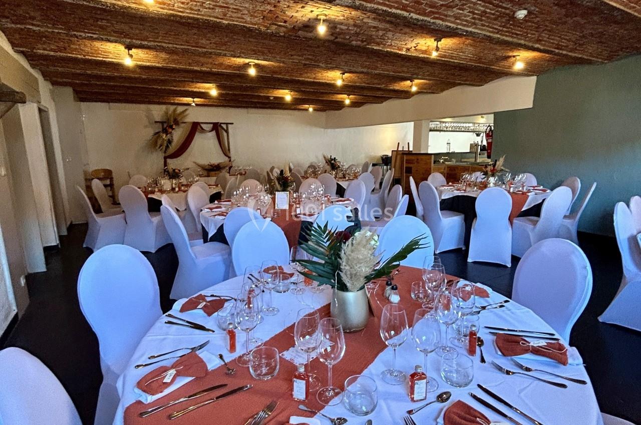 Salle de réception avec tables rondes dressées, nappes blanches, chemins de table orange et plafond en briques apparentes.