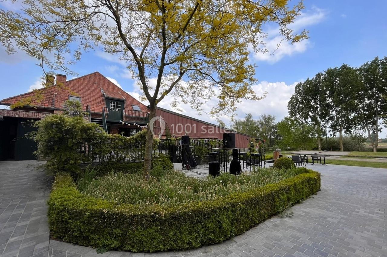 Terrasse extérieure avec mobilier sous un arbre, bordée de haies, près d'un bâtiment en briques rouges et d'un ciel dégagé.