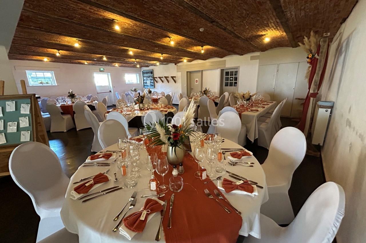 Salle de réception avec tables rondes décorées de nappes blanches, chemins de table orange et compositions florales.