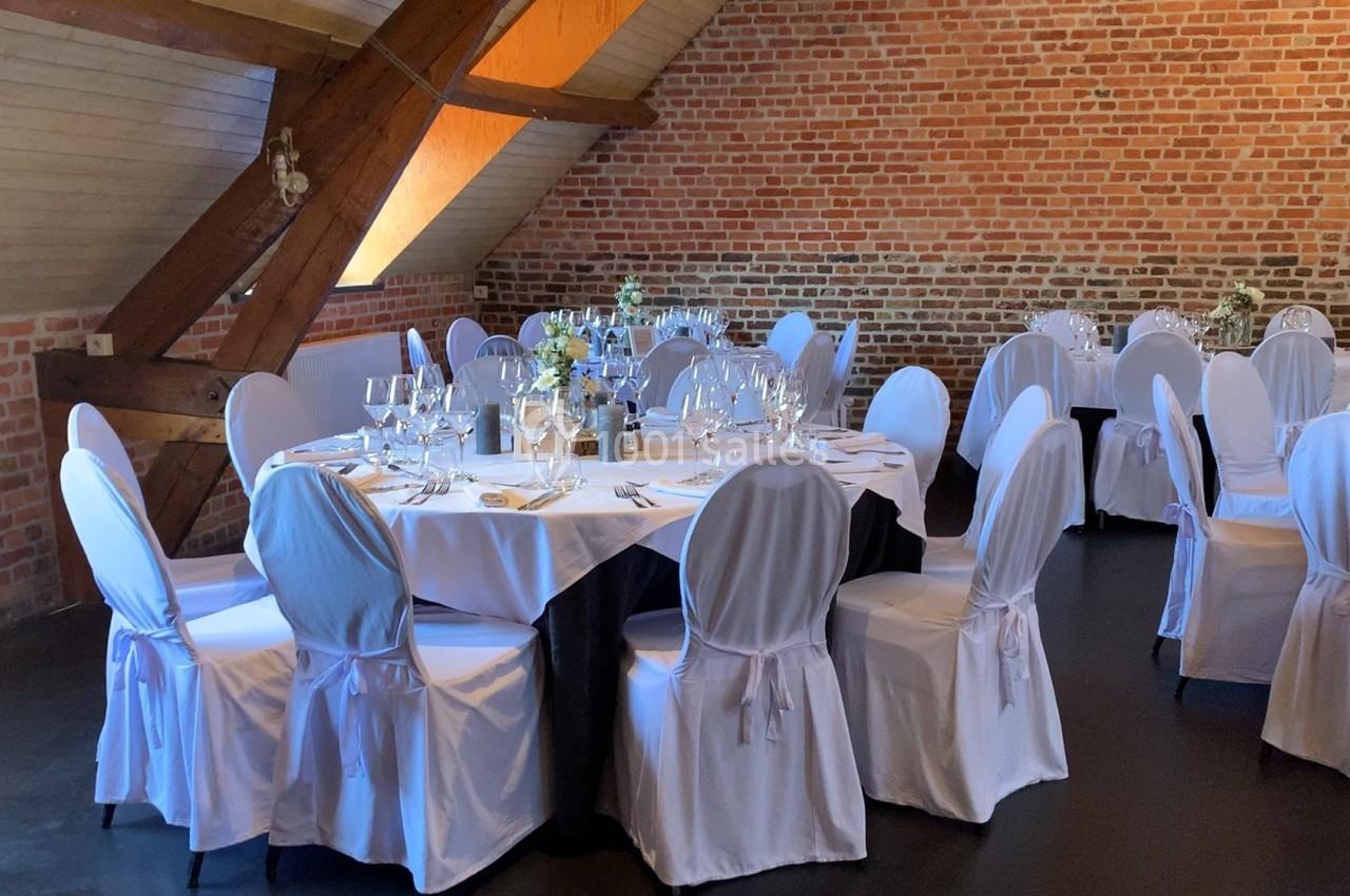 Salle de réception avec tables rondes dressées, nappes blanches et chaises couvertes, dans un décor de briques apparentes.