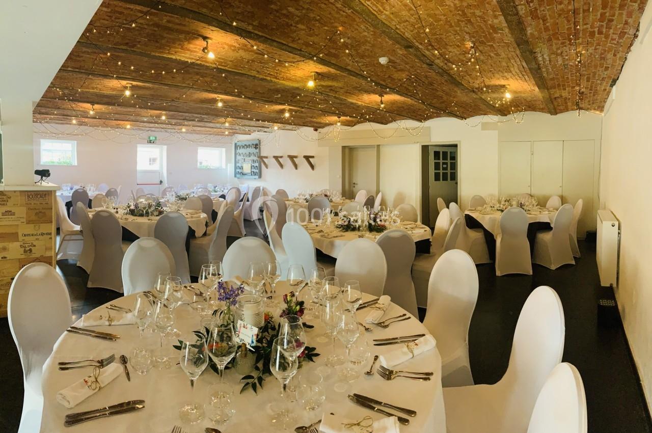 Salle de réception décorée avec des tables rondes dressées, chaises blanches et guirlandes lumineuses au plafond.