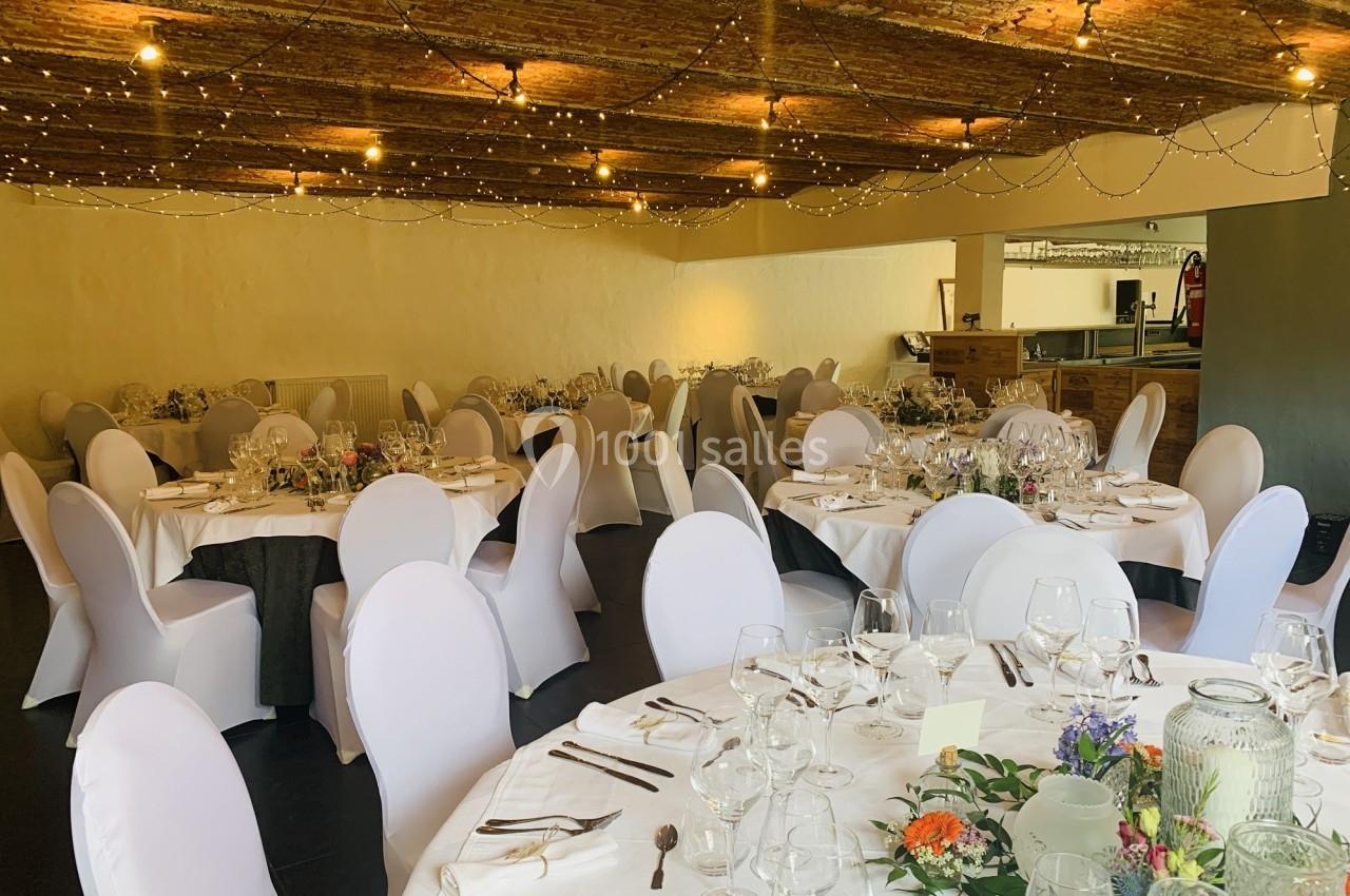 Salle de réception décorée avec des tables rondes dressées, nappes blanches, chaises houssées et guirlandes lumineuses au…