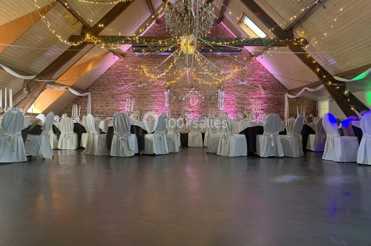 Salle de réception décorée avec des chaises habillées, tables dressées et guirlandes lumineuses suspendues.